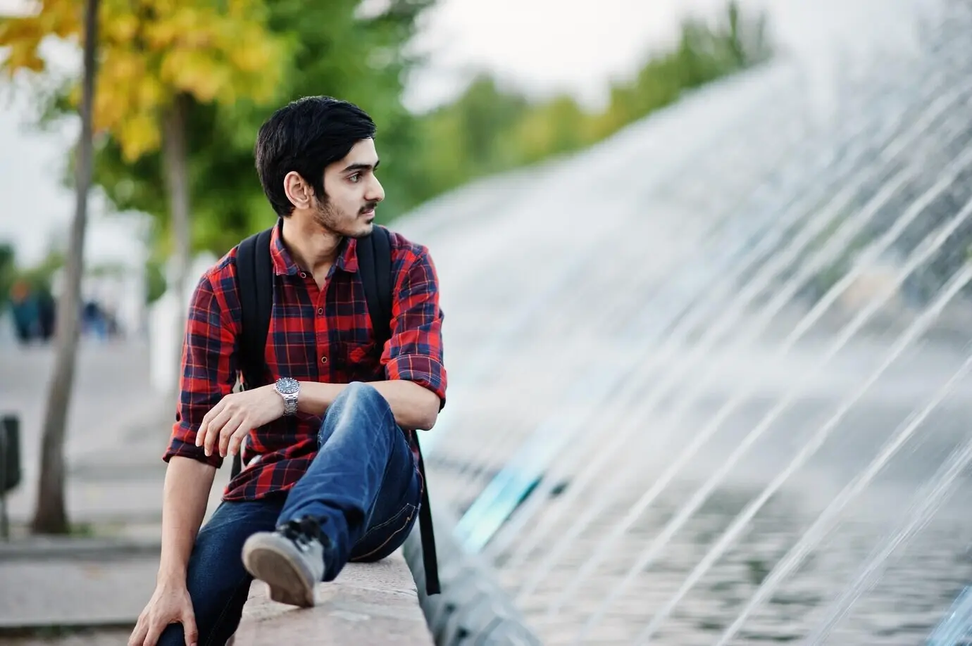 A young Indian male student in a checkered shirt and jeans with a backpack, posing in an evening city setting against the backdrop of fountains.